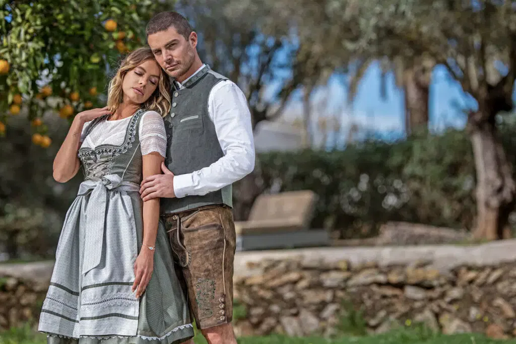 Ein Paar in traditioneller bayerischer Kleidung steht im Freien eng beieinander; die Frau trägt ein Dirndlkleid, der Mann eine Lederhose mit Weste und weißem Hemd. Sie posieren in der Nähe einer Steinmauer und von Bäumen unter einem blauen Himmel.