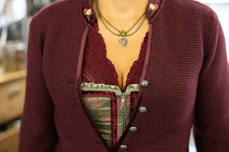 Eine Person trägt eine burgunderfarbene Trachtenjacke mit Zierknöpfen, ein kastanienbraunes Spitzentop und darunter ein grünes Korsett. Zu ihren Accessoires gehören mehrlagige Halsketten, darunter eine mit einem herzförmigen Anhänger.