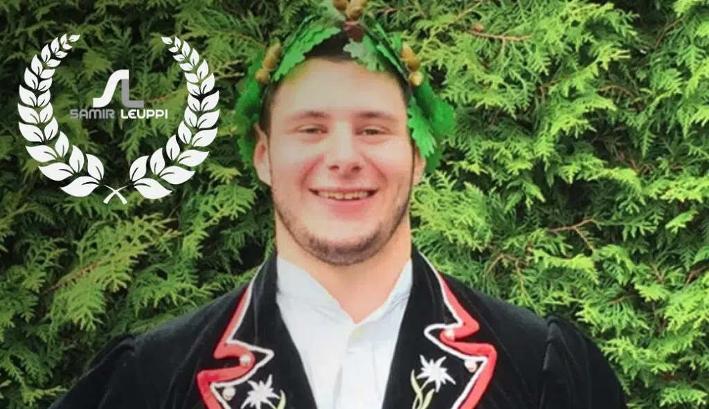 Ein lächelnder Mann in traditioneller Kleidung mit Blattschmuck auf dem Kopf steht vor grünem Laub. Ein weißes Lorbeerkranz-Logo mit dem Text SAMIR LEUPPI und 